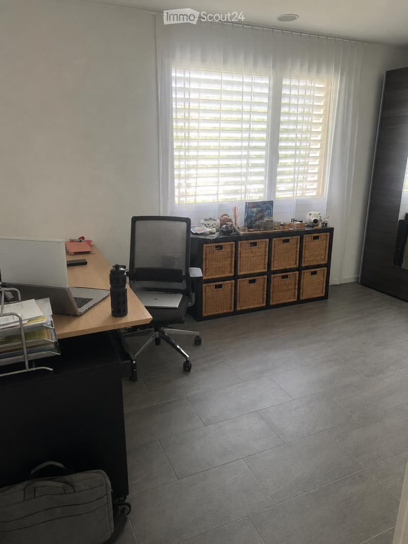 A modern, well-lit office space with a wooden desk, ergonomic chair, laptop, storage unit with woven baskets, and decorative items on top. The room features large windows with blinds and a tiled floor.