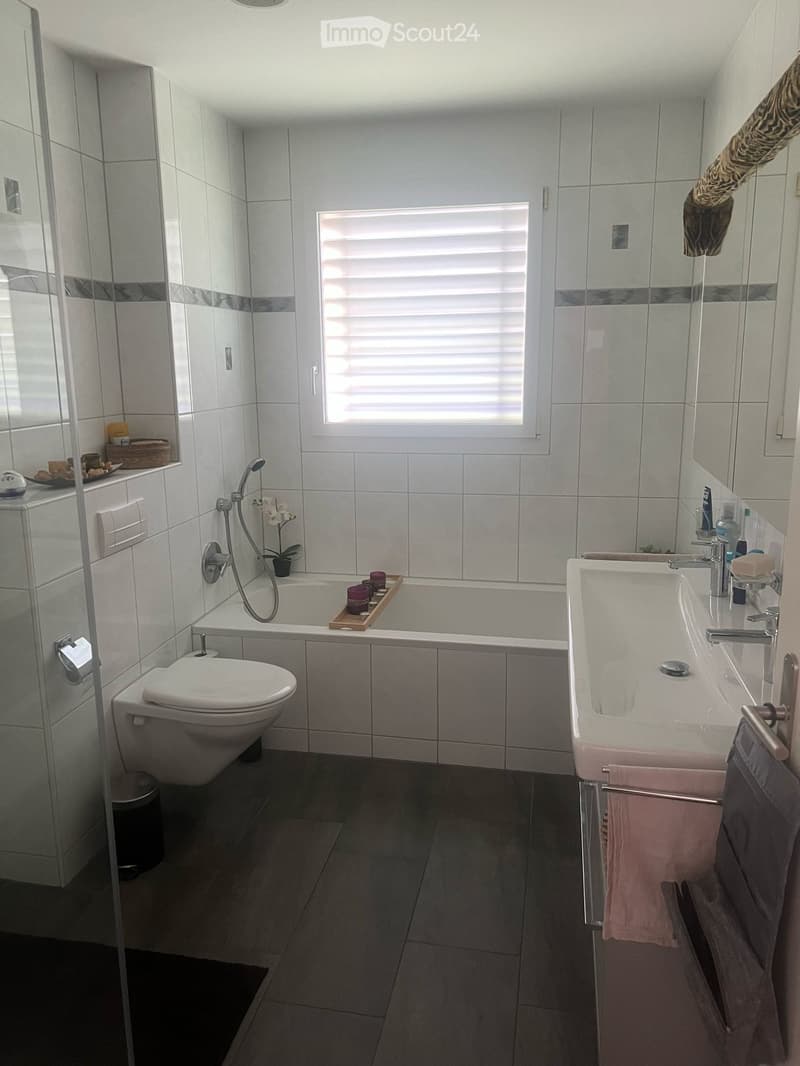 Modern bathroom with white tiles, bathtub, toilet, sink, and a window with blinds. Includes a towel rack with towels, a small flower pot, and a tray with candles.