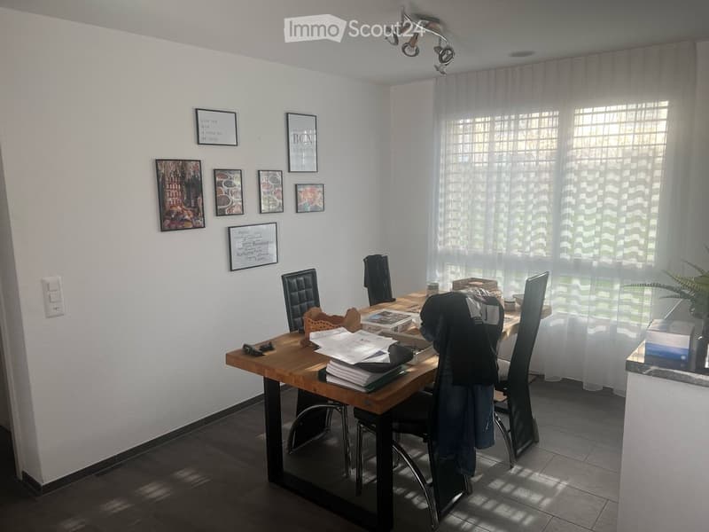 Bright dining area with a wooden table, black chairs, framed pictures on the wall, and a large window with sheer curtains.