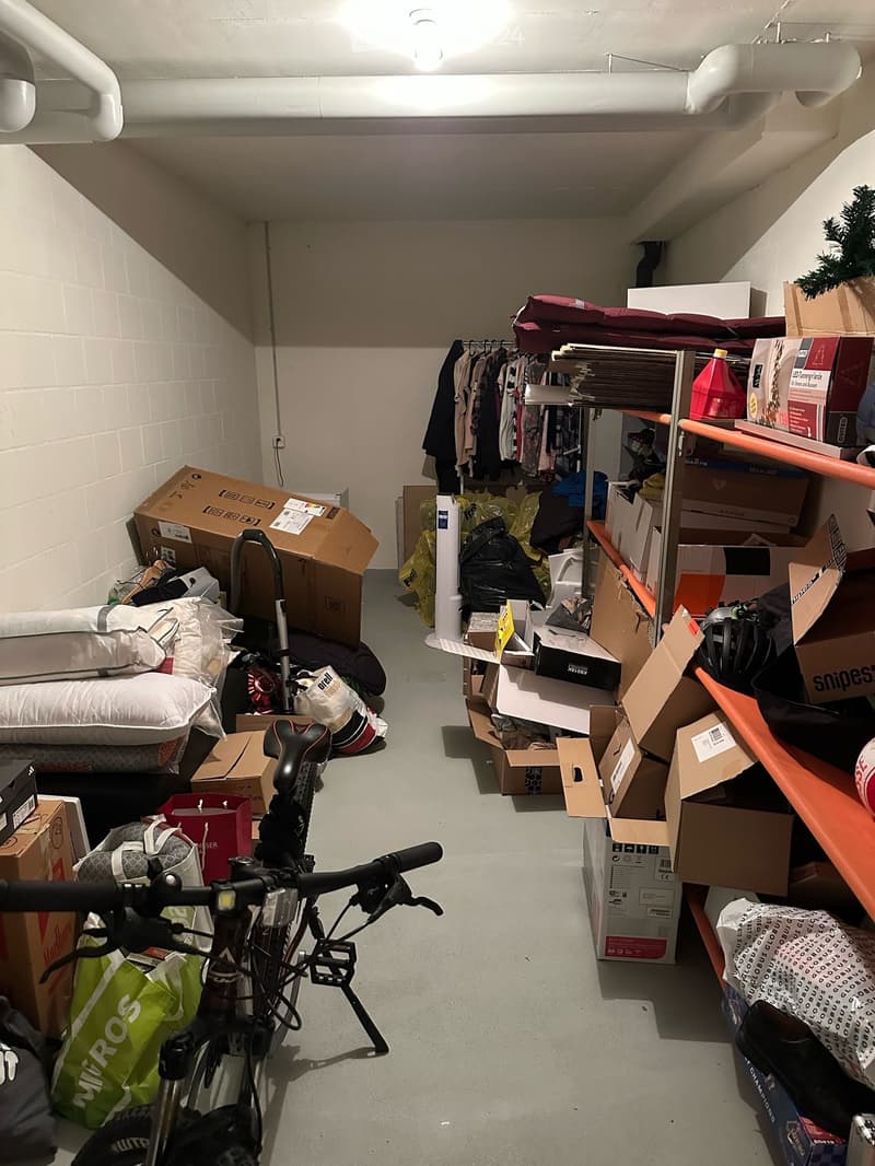 A cluttered storage room with a bicycle, bed, shelves, and various boxes and items scattered around.