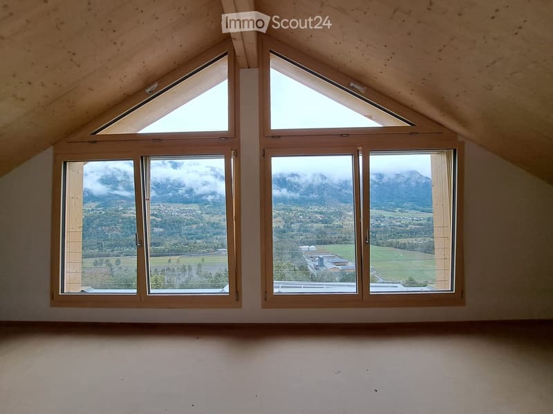An empty attic room with wooden beams, large windows, and a scenic mountain view.