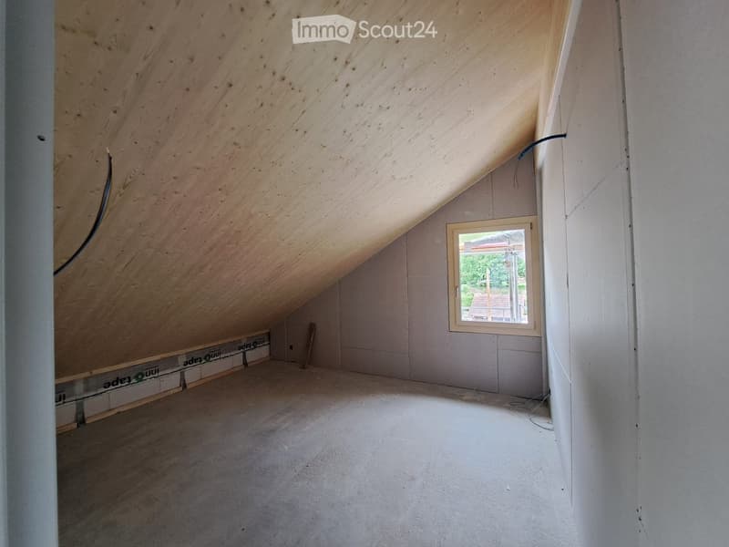 Empty attic space with wooden slanted ceiling, concrete floor, and a single window showing outdoor view.