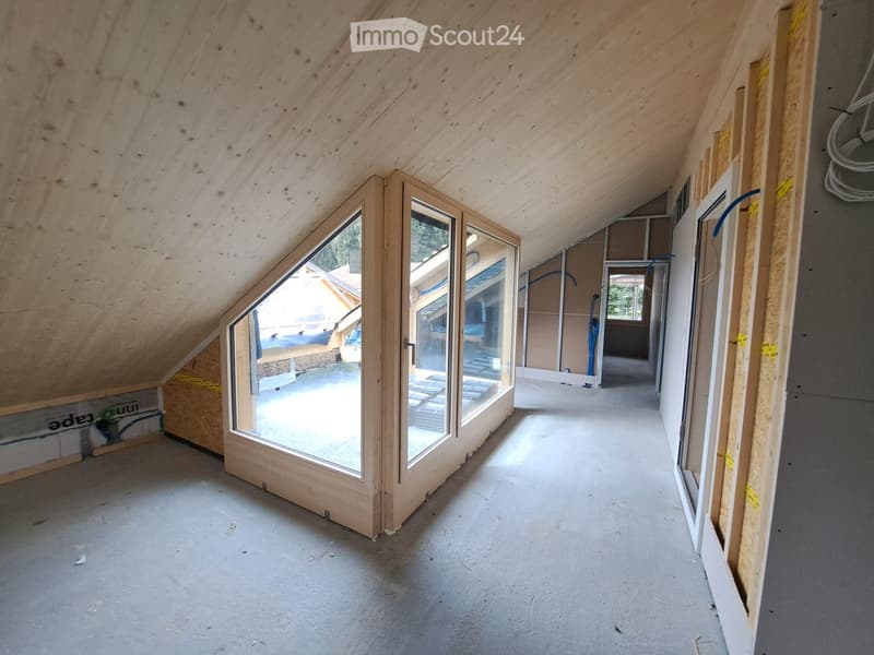 Empty attic space with wooden slanted ceilings, exposed wooden beams, and large windows offering a view of the outdoors. The floor is concrete, and the walls are unfinished with visible insulation and construction materials.