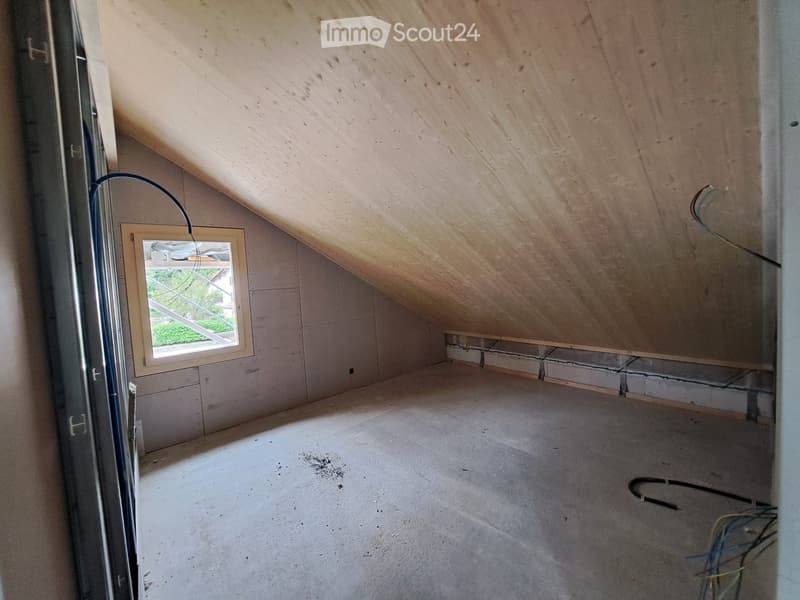 Empty attic space with wooden slanted ceiling, concrete floor, and a single window letting in natural light. Exposed wires and construction materials are visible.