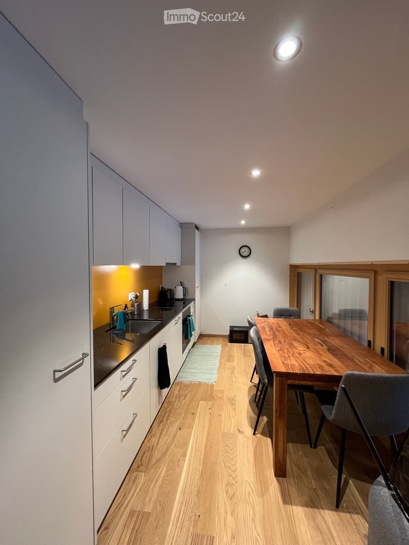 Open-plan kitchen and dining area with light wood flooring, white cabinets, a wooden dining table, and grey chairs.