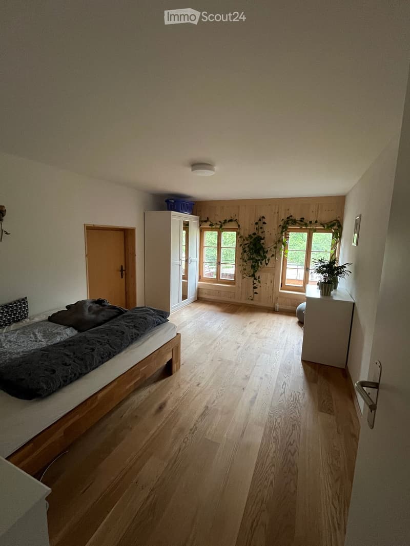 Bright bedroom with wooden flooring, a bed with dark bedding, a wardrobe, a nightstand with a plant, and large windows with wooden frames.