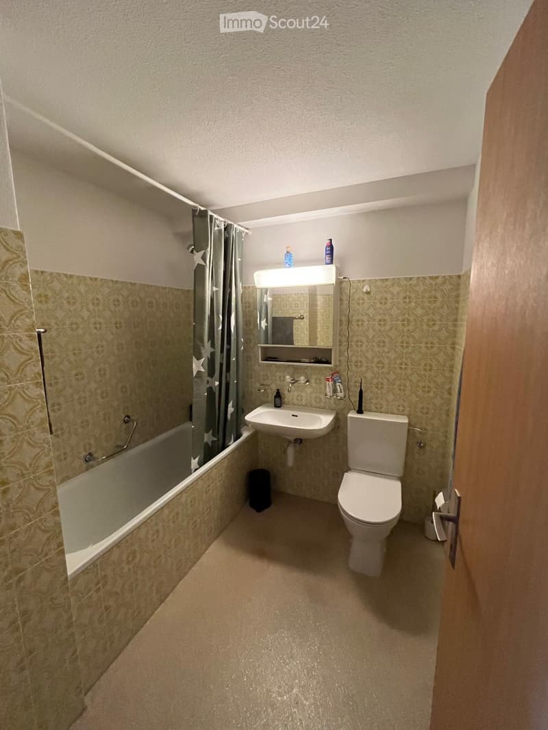 Small bathroom with a toilet, sink, bathtub, and mirror. The walls are tiled with a patterned design, and the floor is carpeted.