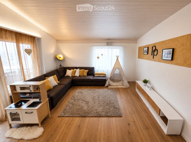 Modern living room with a dark brown sectional sofa, colorful throw pillows, a small kitchenette area, a play tent, and decorative shelves. The room features wooden flooring, large windows with sheer curtains, and warm lighting.