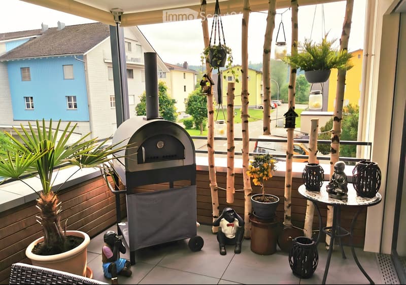 Balcony with a round table, decorative statues, potted plants, a wooden chair, and a wood-burning stove.