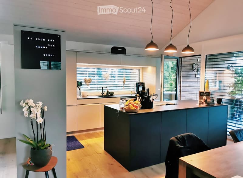 Modern kitchen with a dark blue island, light wood flooring, and large windows. Features include a sink, coffee maker, and decorative elements like a flower vase and framed picture.
