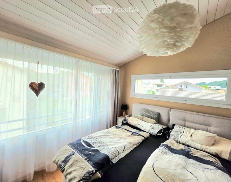Cozy bedroom with a large bed, nightstand, lamp, and large windows with sheer curtains. The room features a slanted wooden ceiling and a decorative heart hanging from the curtain.