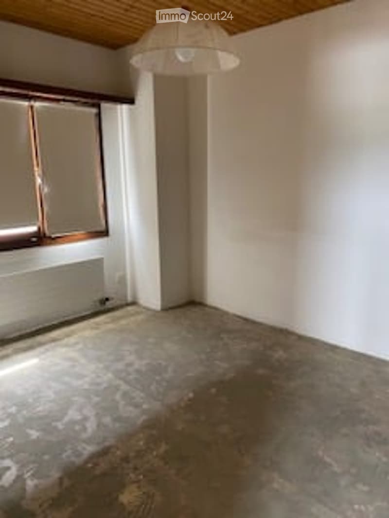 Empty room with white walls, wooden ceiling, and a single window with blinds. The floor shows signs of wear and discoloration.
