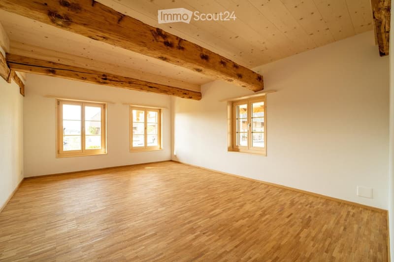 Empty room with wooden floor, exposed wooden beams on the ceiling, and three windows with wooden frames.