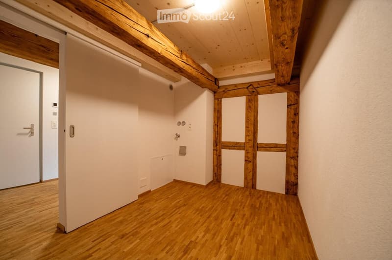Empty hallway with wooden flooring, exposed wooden beams on the ceiling, white walls, and a closed door on the left side.