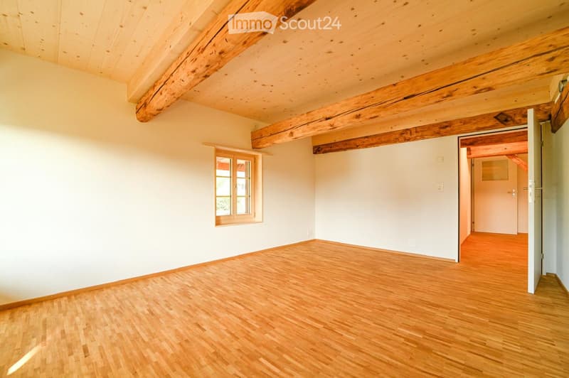 Empty room with wooden flooring, white walls, exposed wooden beams on the ceiling, and a window with wooden frame. An open doorway leads to another room with similar wooden flooring.