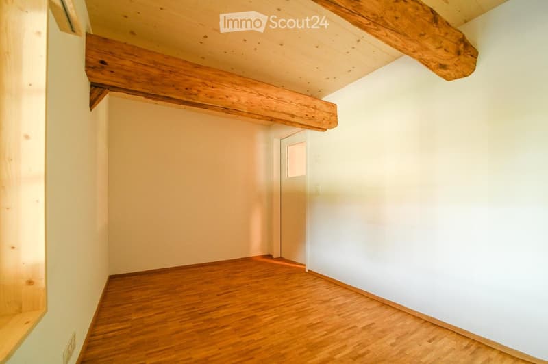 Empty room with wooden floor and exposed wooden beams on the ceiling. The walls are painted white, and there is a door at the far end.