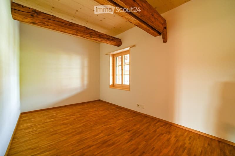 Empty room with wooden flooring, white walls, and exposed wooden beams on the ceiling. A small window with a grid pattern is present on one wall.
