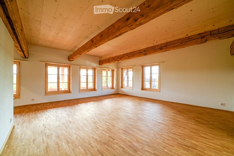 Empty room with wooden floor, exposed wooden beams on the ceiling, and multiple windows with wooden frames.