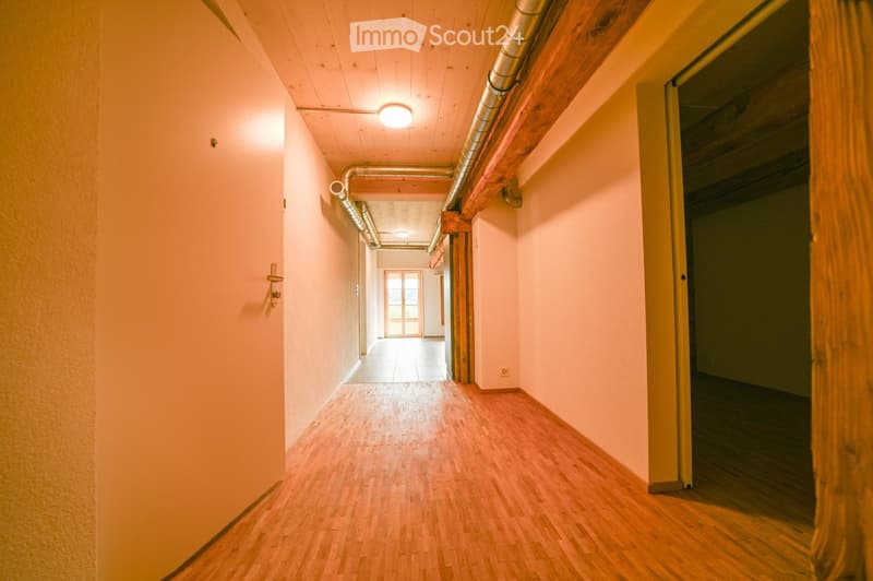 A long, narrow hallway with light pink walls, wooden flooring, and exposed wooden beams. The ceiling has recessed lighting and visible pipes. There are doors on both sides, and the hallway leads to another room with large windows at the end.
