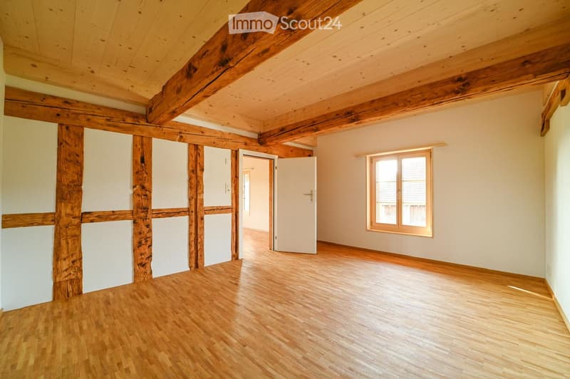 Empty room with wooden floors, exposed wooden beams, white walls, and a single window with wooden frame. No furniture or equipment present.
