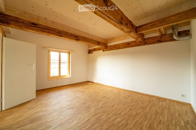 Empty room with wooden floor, exposed wooden beams on the ceiling, white walls, and a single window with wooden frame. No furniture or equipment present.