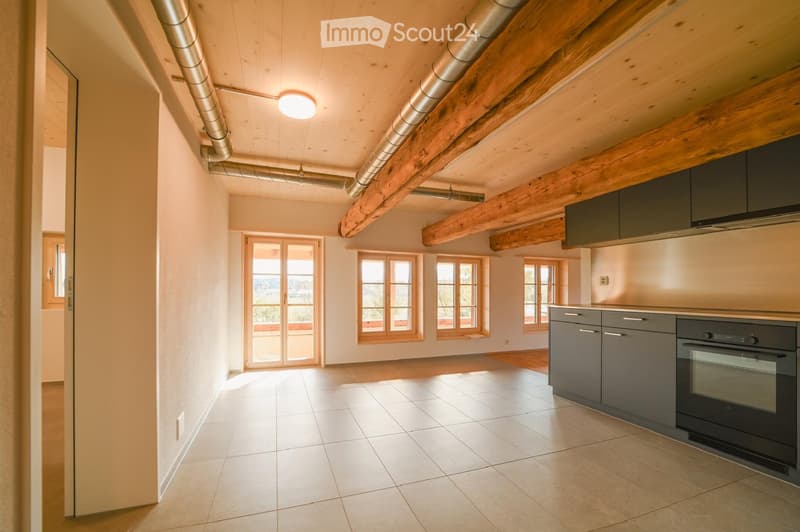 Modern kitchen with grey cabinets, stainless steel oven, and range hood. Exposed wooden beams on the ceiling, large windows, and a sliding glass door leading to a balcony.