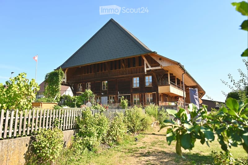 A two-story wooden house with a steeply pitched roof, multiple windows, and a balcony on the upper level. The house is surrounded by greenery and a wooden fence.