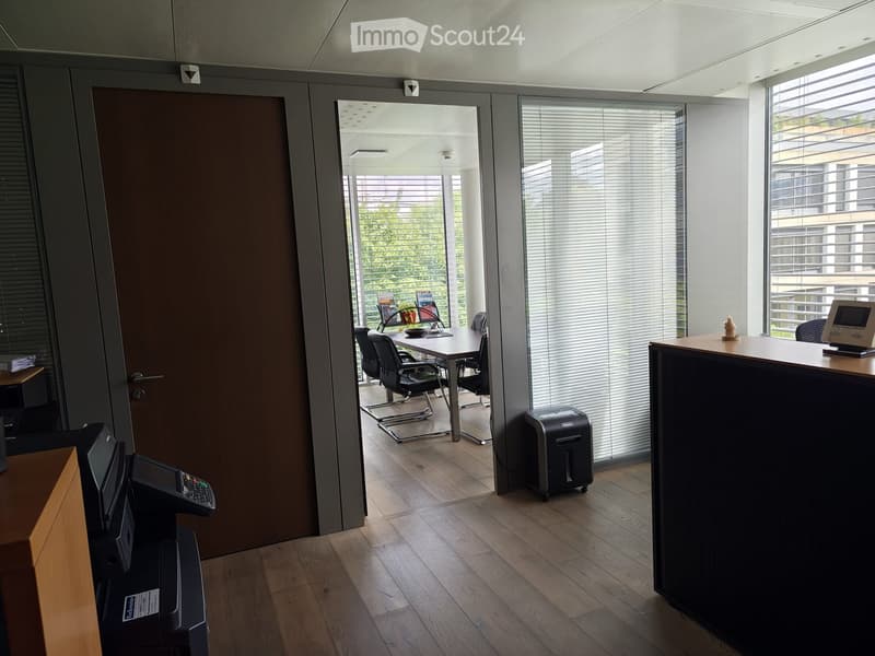 Office space with wooden flooring, large windows with blinds, a desk with chairs, a printer, and a reception area with a screen.