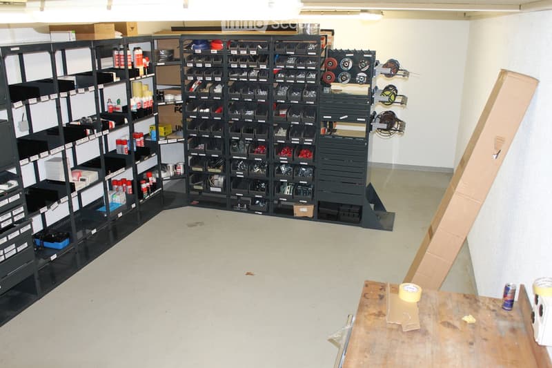 A well-organized workshop or storage area with black shelving units filled with tools and supplies, a wooden workbench with tape and a can, and various boxes and items scattered around.