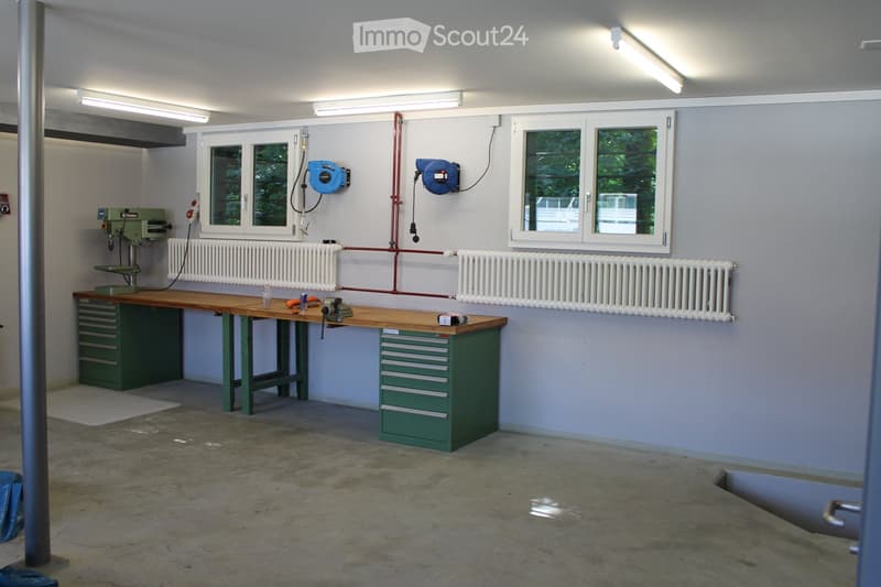 Workshop with a long wooden workbench, green tool cabinet, drill press, radiator, and various tools.