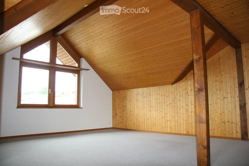 Empty room with wooden ceiling beams, exposed wooden walls, and large windows allowing natural light. The floor is carpeted, and the walls are painted white with wooden paneling on one side.