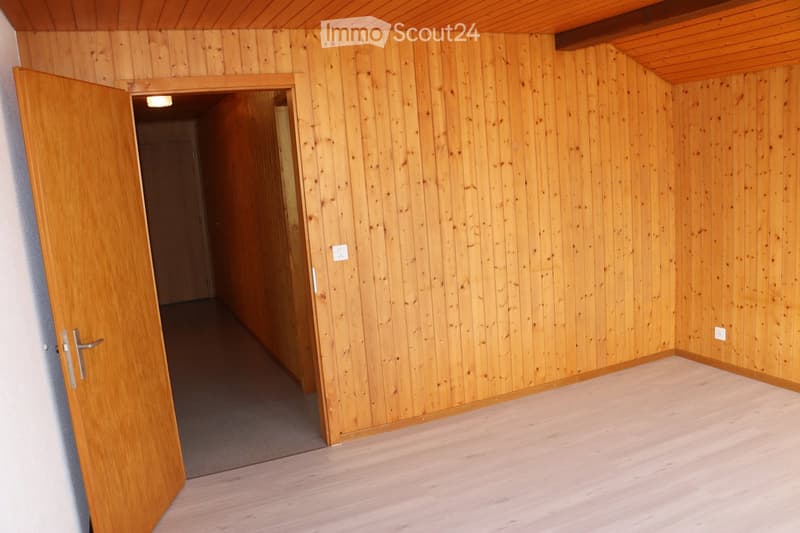 Empty room with wooden walls and floor, featuring an open doorway leading to another space.
