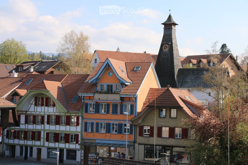 A picturesque village scene with traditional houses featuring colorful facades, red-tiled roofs, and a prominent church tower with a clock in the background.