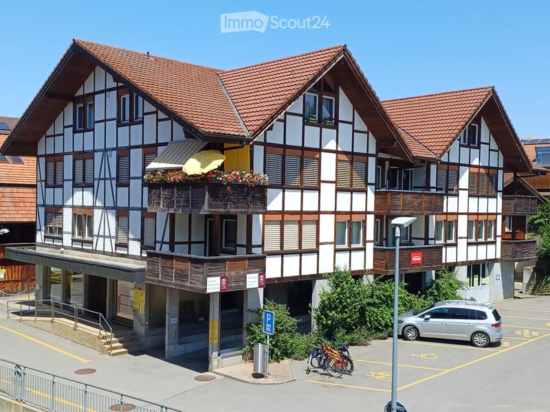 Traditional half-timbered building with red-tiled roof, balconies, and flower boxes. The entrance features a covered walkway and a parking area with a car and bicycles.