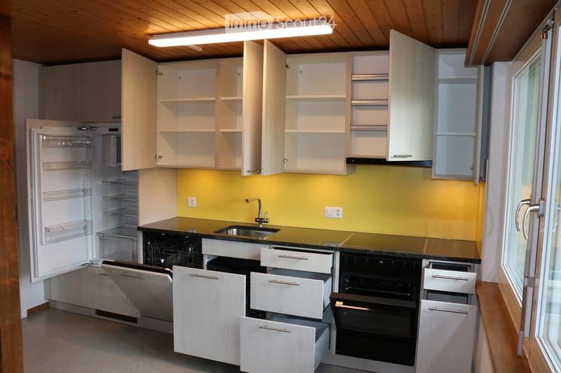 Modern kitchen with white cabinets, black countertops, stainless steel sink, and built-in appliances including a refrigerator and oven. The space features a bright yellow accent wall and ample natural light from a large window.