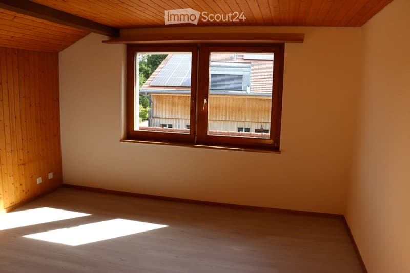 Empty room with wooden walls, wooden floor, and large window offering a view of neighboring buildings and greenery.