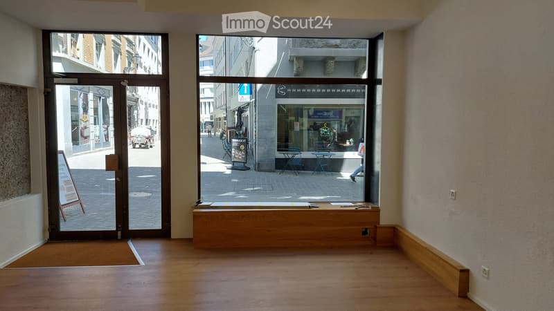 Empty room with wooden flooring, large windows showing a street view, and a wooden bench near the windows.
