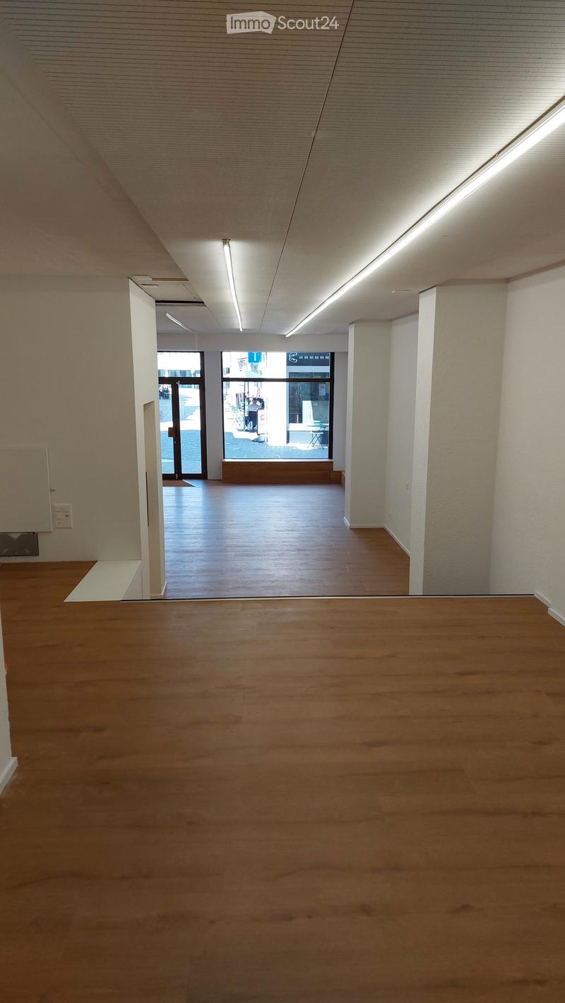 Empty room with wooden flooring, white walls, and large windows leading to an outdoor area. The space is well-lit with recessed lighting.