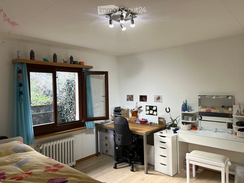 A well-lit bedroom with a bed, desk, chair, mirror, storage unit, and a stool. The room features a large window with blue curtains and a shelf above it. The walls are decorated with pictures and notes.