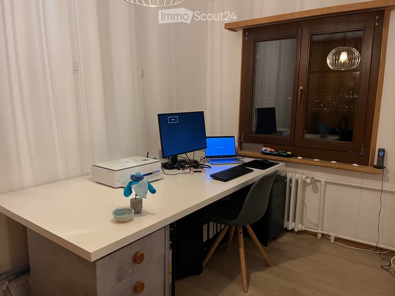 A modern, well-lit workspace with a large desk, laptop, printer, and dual monitors, featuring a comfortable chair and decorative items.