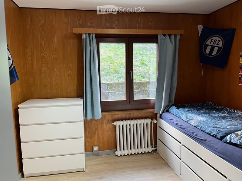Cozy bedroom with wooden walls, a single bed with blue bedding, a white drawer unit, a radiator, and a window with curtains. A flag is displayed on the wall.