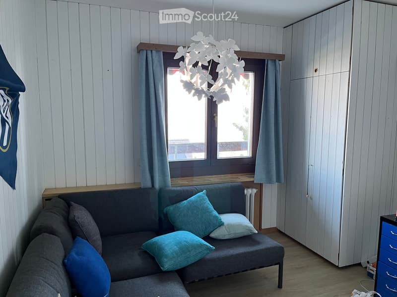A cozy living room with a grey sofa adorned with blue and green pillows, a white floral chandelier, a large window with light blue curtains, and a white cabinet.