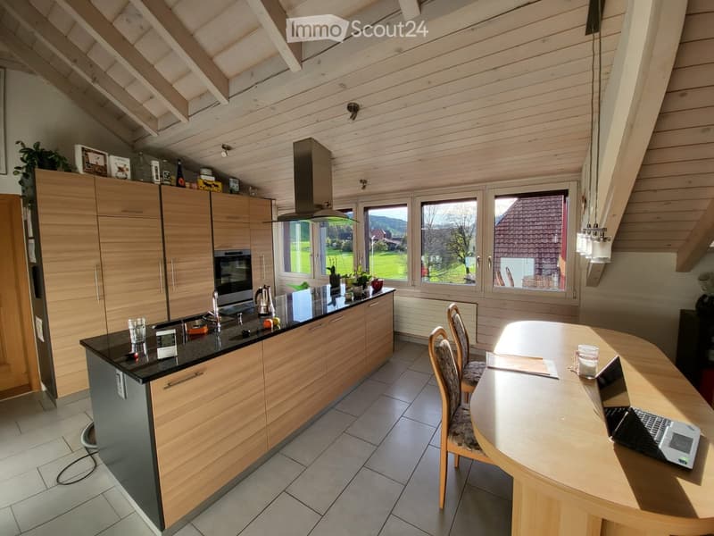 Bright kitchen with wooden cabinets, stainless steel appliances, and a large island with a black countertop. There is a dining area with a round table and chairs, and a view of a green landscape through large windows.