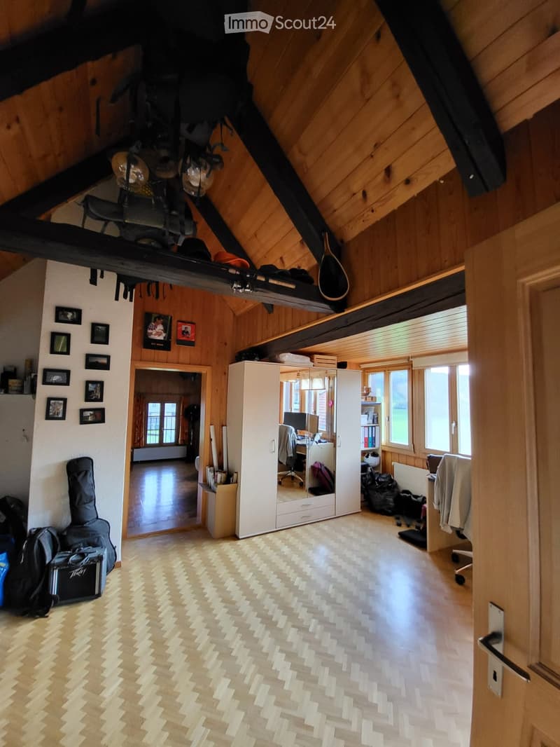 Spacious attic room with wooden beams, herringbone flooring, and large windows. Features a lofted sleeping area, a desk with a laptop, a large white cabinet with a mirror, and various personal items including a guitar and backpacks.