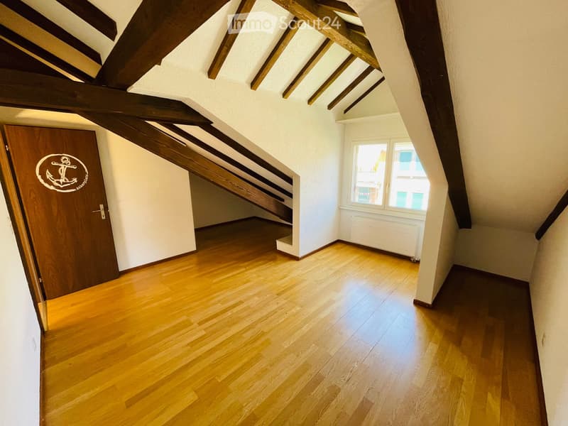 Empty attic space with wooden floors, exposed wooden beams, a staircase leading up, and a window letting in natural light.