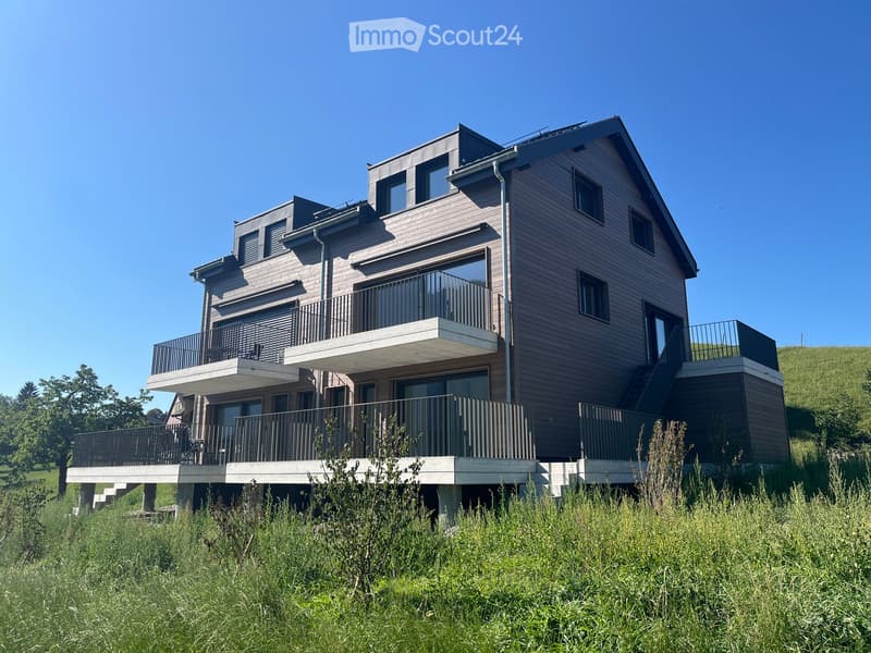 Modern two-story house with wooden siding, multiple balconies, and large windows, situated on a raised ground floor with a grassy slope in front.
