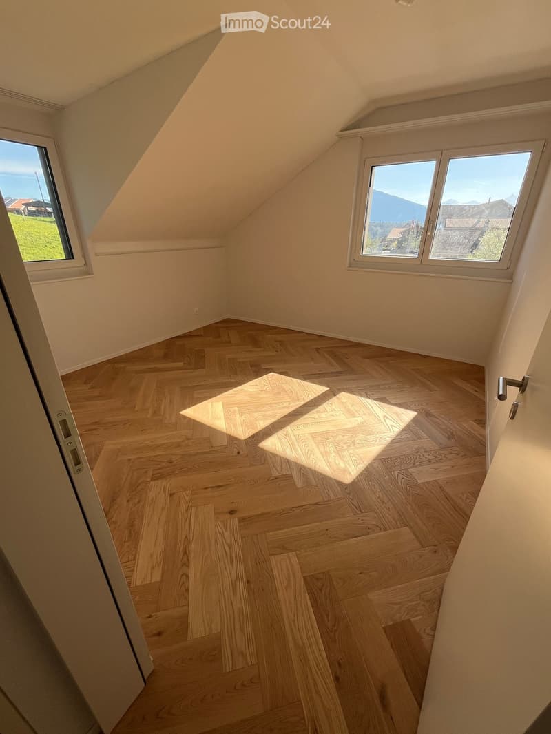 Empty attic room with wooden flooring, large windows, and slanted ceilings.