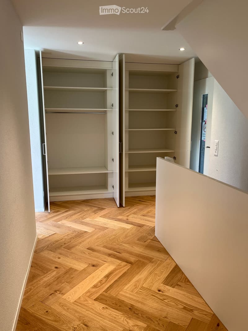 Empty room with wooden herringbone flooring, white built-in closet with open shelves and a hanging rod, and recessed ceiling lighting.