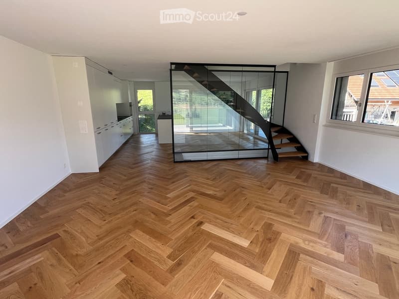 Empty room with herringbone-patterned wooden flooring, white walls, and a modern glass staircase leading to another level. The room is unfurnished, with no visible furniture or equipment.