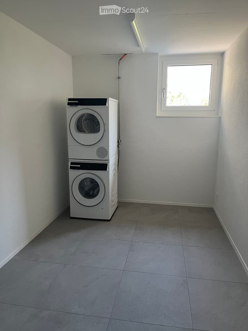 A small, well-lit room with a tiled floor and white walls, featuring a stacked washing machine and dryer unit against one wall and a single window on the opposite wall.